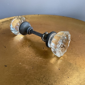 May include: A pair of clear glass door knobs with a metal connecting rod. The knobs have a faceted design and are set against a gold background.