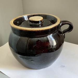 May include: A dark brown glazed ceramic crock with a lid and a single handle. The crock has a light brown rim and a slightly rounded shape.