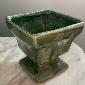 May include: A square, pedestal-style ceramic vase in shades of green. The vase has a glossy finish and a slightly textured surface. The base is square, and the top opening is also square. The overall design is simple and elegant.