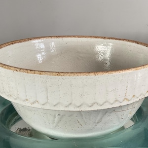 May include: A round ceramic bowl with a speckled off-white glaze and a wide, flat rim. The exterior features a repeating vertical fluted pattern around the upper section, while the rim edge displays a raw, unglazed brown clay texture.