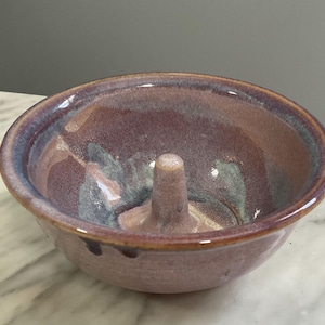 May include: A handmade ceramic apple baker in shades of purple, blue, and brown. The bowl has a central, cylindrical core for holding an apple. The rim is a darker purple, and the interior has a glossy finish.