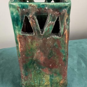May include: A square ceramic vase with a glossy finish, featuring a mottled blend of deep green, brown, and copper tones. The upper section includes triangular cutouts on each side, creating an openwork design with a textured, uneven surface.