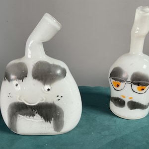 May include: Two white glass vases with hand-painted facial features. The left vase is wide with a thick gray mustache and eyebrows. The right vase is narrower, featuring a gray mustache, orange eyes, and black-rimmed glasses. Both have necks.