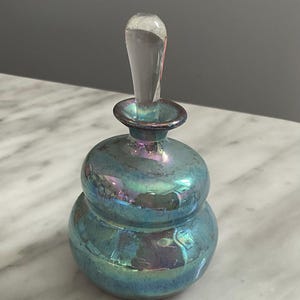 May include: Iridescent teal glass perfume bottle with a clear stopper. The bottle has a bulbous, double-layered body and a slender, clear stopper. The glass reflects shades of blue, green, and purple.