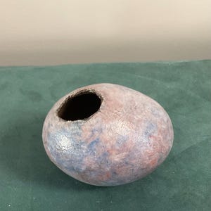 May include: A small, egg-shaped ceramic vessel with a mottled finish in shades of soft pink, light blue, and cream. The object features a single, off-center circular opening at the top with a dark, textured interior rim and a smooth, rounded exterior.