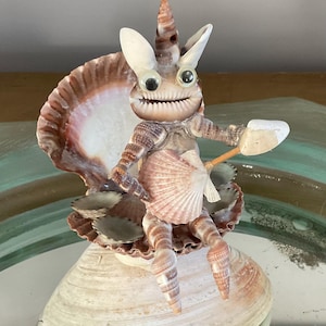 以下が含まれることがあります： クリっとした目、貝殻の体、パドルを持つ生き物を表現した、気まぐれな貝殻彫刻。茶色、ピンク、白の色合いの様々な貝殻で作られており、大きな貝殻の上に設置されています。