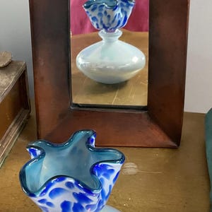 May include: A decorative glass vase with a ruffled rim and a blue and white spotted pattern. The vase sits on a round, iridescent base. A wooden framed mirror reflects the vase.