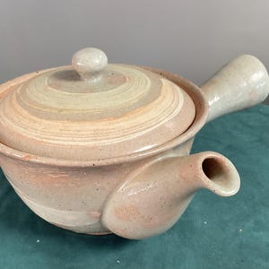 May include: A ceramic teapot with a side handle and a matching lid. The teapot features a speckled, light beige and soft peach glaze with subtle horizontal banding on the lid. It has a short, wide spout and a rounded body shape.