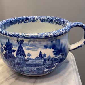 May include: A blue and white ceramic chamber pot with a handle. The pot features a landscape scene in blue and white, with a decorative rim. The handle is also blue and white.