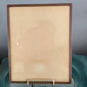 May include: A rectangular brown picture frame with a light brown interior. The frame is supported by a silver-colored metal stand. The frame is empty and ready for a photo or artwork.