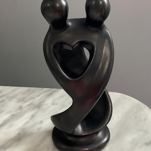 May include: A dark brown sculpture depicting two figures embracing, with a heart-shaped cutout in the center. The sculpture has a smooth, polished finish and sits on a round base. The figures' heads are rounded, and the overall design is abstract.