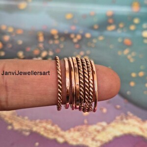Op de afbeelding: Een verzameling koperkleurige ringen, sommige met een gedraaid touwontwerp, andere glad en plat. De ringen zijn op een vinger gestapeld en tonen hun verschillende texturen en stijlen. De ringen hebben een warme, metallic tint.