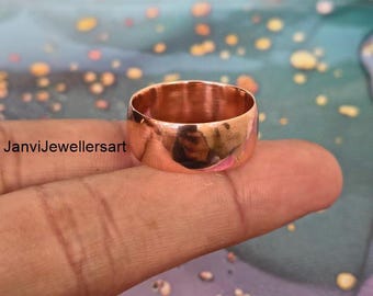 May include: A polished copper ring with a wide band is displayed. The ring reflects the surrounding environment, showcasing its smooth, shiny surface. The ring is held on a finger, with the text "JanviJewellersart" visible in the background.