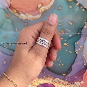 May include: A silver ring with a leaf design on the middle band is worn on a finger. The ring has a smooth, polished finish. The background is a colorful abstract pattern.