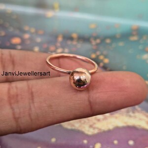 Könnte beinhalten: Ein roségoldener Ring mit kugelförmigem Design. Der Ring hat ein dünnes Band und eine polierte Kugel. Der Ring wird auf einem Finger präsentiert und zeigt sein schlichtes, elegantes Design. Der Text "JanviJewellersart" ist sichtbar.