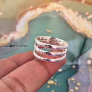 May include: A silver ring with three bands is held between fingers. The ring is shiny and reflective, with a smooth surface. The ring is a piece of jewelry.