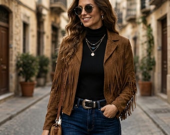 May include: A brown suede jacket with long fringe detailing on the sleeves, worn over a black turtleneck sweater and dark blue denim jeans. The outfit includes a black leather belt with a silver buckle, layered gold necklaces, and round sunglasses.