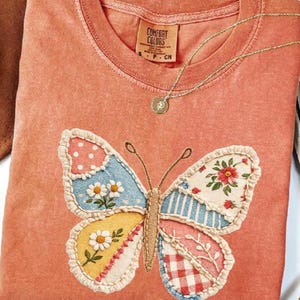 May include: Coral-colored t-shirt with a large, embroidered butterfly applique. The butterfly features patchwork wings with floral, polka dot, and striped patterns. A gold necklace with a pendant rests on the shirt. The tag reads "Comfort Colors".