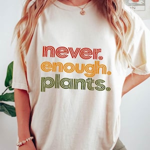 May include: Cream-colored t-shirt with the text "NEVER. ENOUGH. PLANTS." in a retro font. The text is layered in red, orange, and green. The shirt is a relaxed fit.