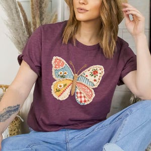 May include: A maroon t-shirt featuring a colorful butterfly applique. The butterfly design incorporates various patterns, including floral, polka dot, and plaid, in shades of blue, yellow, and red. The shirt is paired with blue jeans.