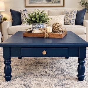May include: A square navy blue coffee table with turned legs and a single drawer featuring a round gold knob. On top sits a rectangular wooden tray containing a blue and white patterned planter with a green plant, a candle, and a decorative orb.