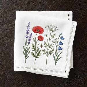 May include: White linen napkin featuring an embroidered floral design. The design includes lavender, red poppies, white Queen Anne's lace, and bluebell flowers. The napkin has a decorative cutwork border.