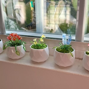 May include: Five miniature potted plants featuring beaded flowers in red, orange, yellow, blue, and pink. Each plant is set in a white, geometric, faceted ceramic pot filled with green moss. The flowers are crafted from small glass beads.