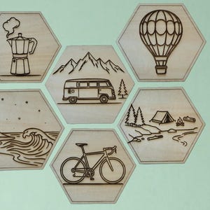 May include: A set of six light-coloured wooden hexagonal wall plaques featuring laser-engraved illustrations of a moka pot, a camper van with mountains, a hot air balloon, a wave with stars, a bicycle, and a campsite with a tent and campfire.
