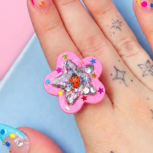 May include: A chunky pink flower-shaped ring featuring a central star-shaped cavity filled with clear resin, silver glitter, and a small red ladybug charm. The ring is worn on a finger with long, pointed nails decorated with colorful dots and rhinestones.