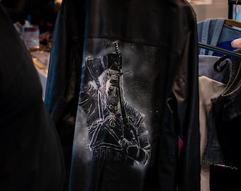 May include: A black leather jacket featuring a detailed white airbrushed illustration on the back. The artwork depicts a warrior figure with long hair holding a sword over their shoulder, surrounded by a subtle stippled effect and light highlights.