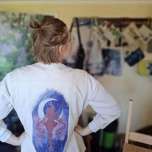 Puede incluir: Chaqueta vaquera blanca con un diseño pintado a mano en la espalda. El diseño presenta la silueta de un personaje con pelo largo y suelto, una luna creciente y un cielo estrellado en tonos morados y azules.