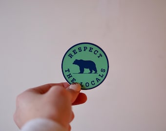 May include: A round sticker with a green background and a blue border. The sticker features a blue bear silhouette and the text "RESPECT THE LOCALS" in blue. The sticker is held by a hand against a white background.