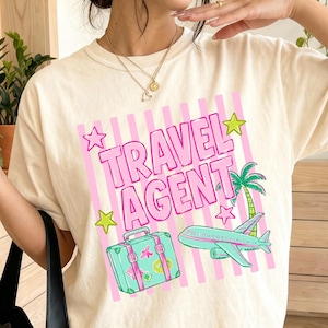 May include: An ivory short-sleeve t-shirt featuring a graphic print with pink vertical stripes and the text TRAVEL AGENT in pink. The design includes a teal suitcase, a teal airplane, a green palm tree, and yellow stars on the front.