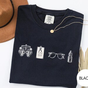 May include: A black short-sleeved t-shirt featuring a white graphic print of an optometry phoropter, a Snellen eye chart, a pair of clear-framed glasses, and a small dropper bottle. The shirt has a Comfort Colors brand label at the inner neckline.