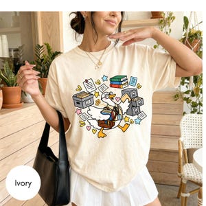 Könnte beinhalten: Ein elfenbeinfarbenes T-Shirt mit einer bunten Grafik einer Cartoon-Gans, die eine Brille und ein Schlüsselband trägt sowie eine braune Umhängetasche und Bücher hält. Das Motiv zeigt Bücherstapel, Aktenschränke und verstreute Papiere.