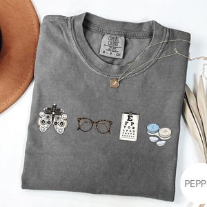 May include: A charcoal grey t-shirt featuring a graphic print of optometry equipment including a phoropter, tortoise shell glasses, an eye chart with the letters E, F, P, T, O, Z, L, P, E, D, and a contact lens case with two contact lenses.