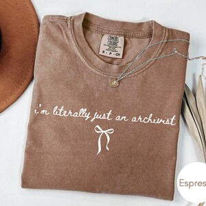 May include: An espresso-coloured short-sleeve t-shirt with a crew neckline. The shirt features the white text "i'm literally just an archivist" printed in a cursive font above a simple white line-drawn bow graphic on the centre of the chest.