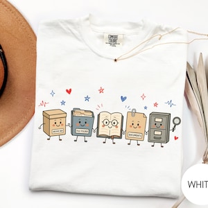 May include: A white t-shirt featuring a graphic of five anthropomorphic office items holding hands: an archive box, a file folder, an open book with glasses, a document sheet with a paperclip, and a filing cabinet holding a magnifying glass.