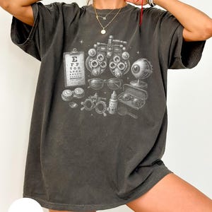 May include: A charcoal grey short-sleeve t-shirt featuring a white graphic print of optometry equipment, including a phoropter, an eye chart, a human eye model, glasses, a contact lens case, and a bottle labelled eye drops, with small star accents.