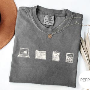 May include: A charcoal grey short-sleeved t-shirt featuring a graphic print of a laptop with a bar chart, an open ledger with a line graph, a clipboard with an upward trend, and a calculator. The shirt has a Comfort Colors brand label at the neck.