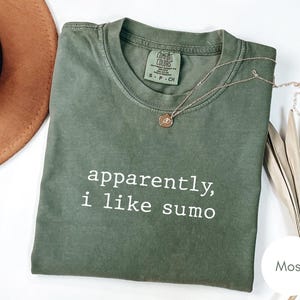 May include: A moss green t-shirt with the text "apparently, i like sumo" in white. A gold necklace with a pendant rests on the shirt. A brown hat and dried plant are in the background.