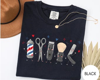 Puede incluir: Una camiseta negra de manga corta con un gráfico de cinco herramientas de barbero antropomórficas tomadas de la mano: un poste de barbero, tijeras, cortapelos, una brocha de afeitar y una navaja, decorada con corazones y estrellas rojas y azules.