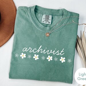 Könnte beinhalten: Ein hellgrünes Kurzarm-T-Shirt mit Rundhalsausschnitt. Das Shirt zeigt das Wort archivist in weißer Schreibschrift über einer Reihe von weißen und gelben Blumenillustrationen. Das Etikett im Kragen zeigt Comfort Colors S P CH.