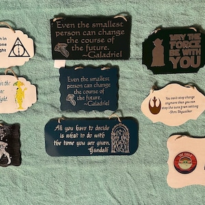 May include: A collection of wooden signs with quotes from popular movies and books. The signs feature quotes like "May the Force be with you" and "Precious Cargo" with character illustrations. The signs are various shapes and colors.