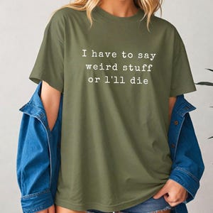 Pode incluir: Camiseta verde-oliva com o texto branco: "I have to say weird stuff or I'll die". Uma jaqueta jeans azul é usada sobre a camiseta. A camiseta é de estilo casual.