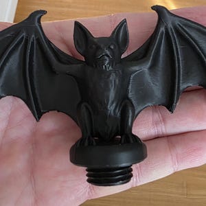 Close-up of a black gothic bat topper held in a hand, featuring detailed wings spread wide, sculpted bat face and body, and a threaded base designed to screw securely into the lid of a gothic storage canister.