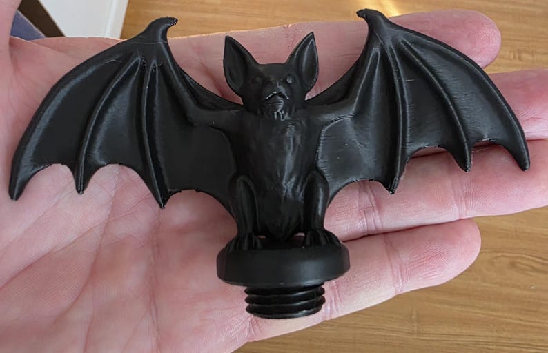 Close-up of a black gothic bat topper held in a hand, featuring detailed wings spread wide, sculpted bat face and body, and a threaded base designed to screw securely into the lid of a gothic storage canister.
