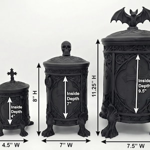 May include: Three black cylindrical lidded containers with gothic detailing, cross motifs, and claw feet. The smallest is 5.5 inches high and 4.5 inches wide with 4-inch depth. The medium is 8 inches high and 7 inches wide with 7-inch depth. The largest is 11.25 inches high and 7.5 inches wide with 9.5-inch depth.