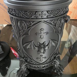 Open black gothic bat canister without lid, showcasing deep interior storage, ornate medieval scrollwork, raised cross accents, shield panel with bat emblem, side ring handles, and clawed feet, photographed on a glass table to highlight texture and capacity.