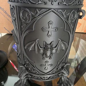 Close-up of gothic bat canister showing ornate carved details, raised bat emblem, cross accents, and clawed feet, highlighting dark gothic craftsmanship and textured matte black finish.